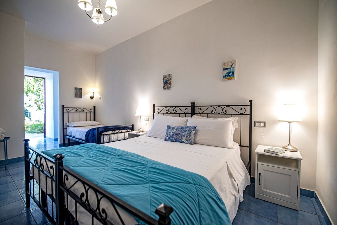A bright, well-lit bedroom with two wrought iron beds featuring white linens and turquoise accents, side tables with lamps, and access to an outdoor patio area.