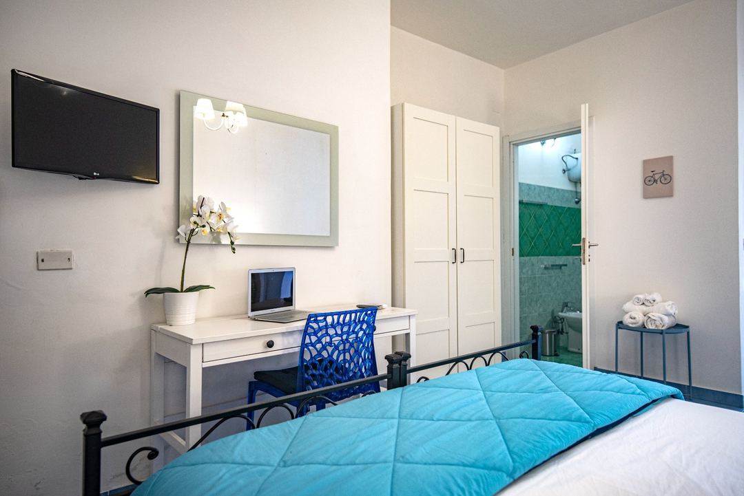 A bright bedroom at Sonnino B&B featuring a blue quilted comforter on a wrought iron bed, white desk with work area, and an ensuite bathroom visible through glass doors.