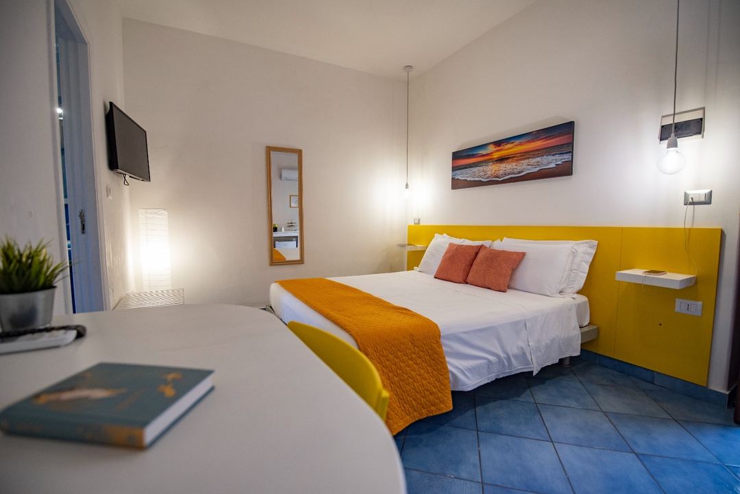 Bright modern bedroom with a yellow headboard, white linens, orange accent pillows, and blue tiled flooring at Sonnino B&B.