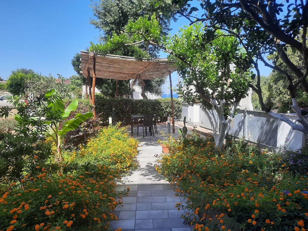A charming garden pathway lined with vibrant orange marigolds leads to a wooden pergola with seating area and Mediterranean architecture in the background.