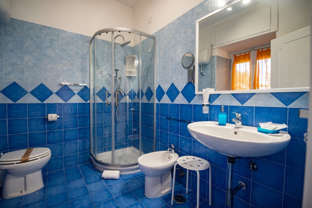A bright blue-tiled bathroom featuring a curved glass shower enclosure, pedestal sink, bidet, and toilet with modern fixtures and natural light from a window.