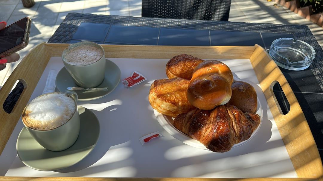A breakfast tray featuring two cappuccinos, fresh croissants, and pastries served on a wooden tray in bright natural sunlight.