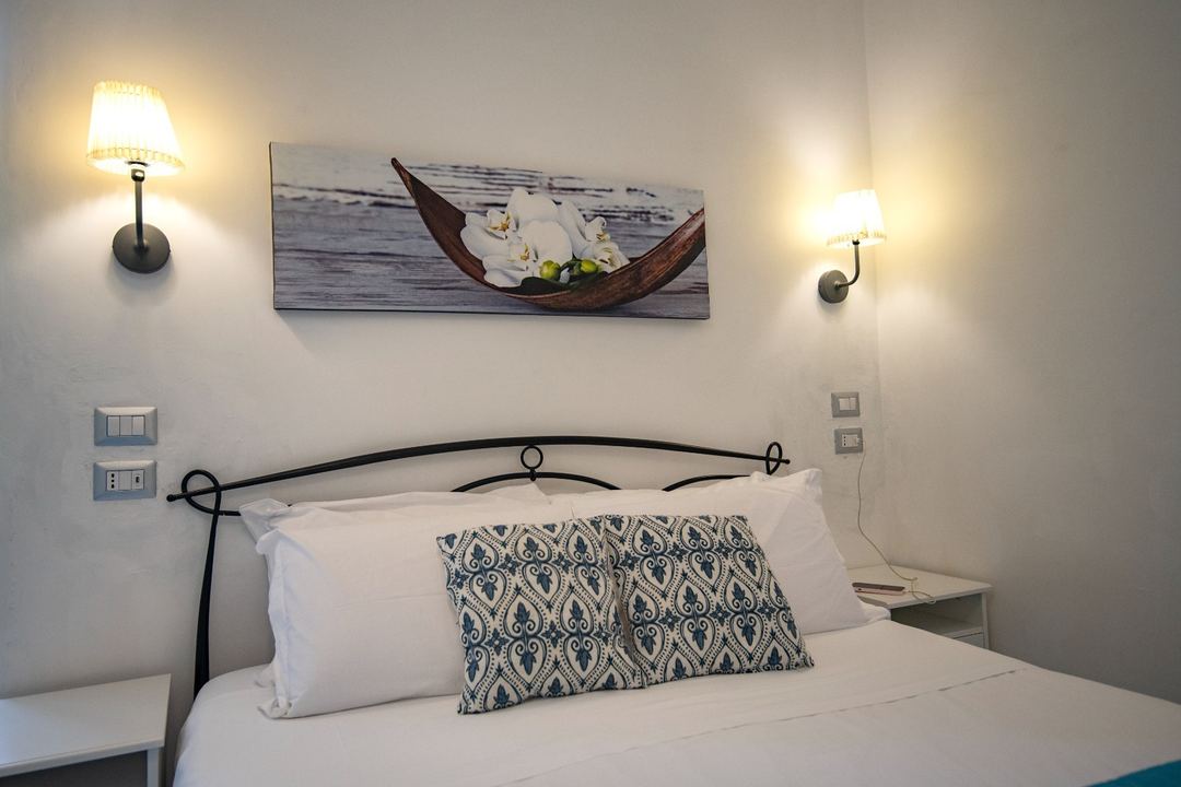 A bright bedroom with a black metal bed frame, white pillows, patterned cushion, and decorative wall sconces flanking a boat-themed artwork.